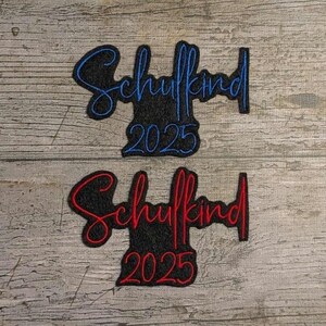 May include: Two embroidered patches with the word "Schulkind" and the year "2025". One patch has blue lettering on a black background, and the other has red lettering on a black background. The patches are on a weathered wood surface.