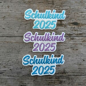 May include: Three embroidered patches with the German word "Schulkind" and the year "2025". The patches are in blue and purple, with white borders, set against a rustic wooden background.