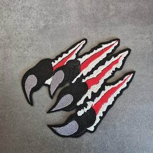 Könnte beinhalten: Gestickte Krallenaufnäher. Jeder Aufnäher zeigt eine schwarze Kralle mit grauen Akzenten und rot-weißen Streifen. Diese Aufnäher sind zum Aufnähen oder Aufbügeln auf Stoff konzipiert, ideal zum Anpassen von Kleidung oder Accessoires.