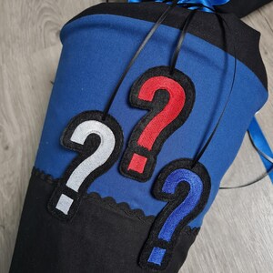 Puede incluir: Una bolsa escolar negra, azul y blanca con tres apliques de signo de interrogación en blanco, rojo y azul. La bolsa tiene un cierre de cordón azul y una cinta azul. La bolsa está hecha de fieltro.