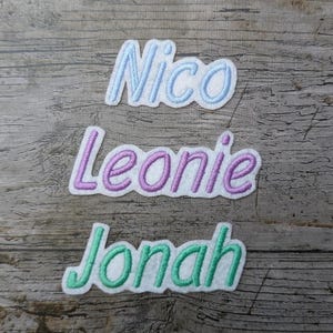 May include: Three embroidered name patches: "Nico" in light blue, "Leonie" in purple, and "Jonah" in green. Each name is stitched with a white outline, set against a weathered wood background. These patches are ideal for personalizing items.