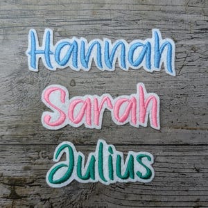 May include: Three embroidered name patches: "Hannah" in blue, "Sarah" in pink, and "Julius" in teal. Each name is stitched in a cursive font with a white border. The patches are displayed on a weathered wooden surface.