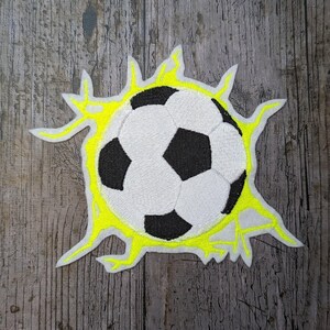 Könnte beinhalten: Gestickter Aufnäher mit einem Fußball-Design, mit schwarz-weißen Paneelen, umgeben von einem gelben Burst-Effekt. Der Aufnäher befindet sich auf einer Holzoberfläche, ideal für Kleidung oder Accessoires.