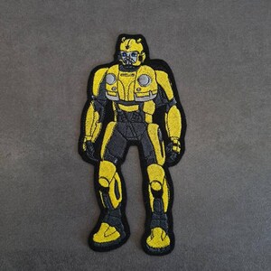 Könnte beinhalten: Gestickter Aufnäher mit einem gelb-schwarzen Robotercharakter. Der Roboter hat ein detailliertes Design mit gelben Akzenten auf einer schwarzen Kontur. Der Aufnäher ist wahrscheinlich für Kleidung oder Accessoires und ist ein Popkultur-Artikel.