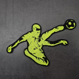 Peut inclure: Écusson brodé vert fluo et noir représentant un joueur de football en train de frapper dans un ballon. L'écusson a la forme d'un joueur de football en plein coup de pied, le ballon étant positionné près du pied. L'écusson a une bordure noire.