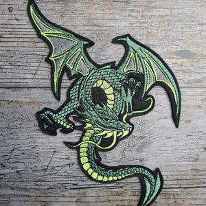 Könnte beinhalten: Gestickter Aufnäher mit einem grünen Drachen mit detaillierten Schuppen, Flügeln und einem langen Schwanz. Der Drache hat eine schwarze Umrandung und limettengrüne Akzente. Der Aufnäher ist zum Aufnähen oder Aufbügeln auf Stoff gedacht.