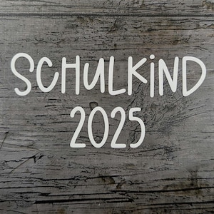 May include: White text on a gray wooden background reads "SCHULKIND 2025". The text is in a handwritten style. The background has a wood grain texture.