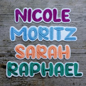 May include: Embroidered name patches in purple, blue, orange, and green. The names "Nicole", "Moritz", "Sarah", and "Raphael" are displayed on a wooden background. These patches are ideal for personalizing clothing or accessories.