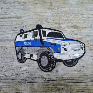 Peut inclure: Écusson brodé représentant un véhicule blindé bleu, blanc et gris avec le mot "POLIZEI" en bleu. Le patch a de gros pneus noirs et un tuba noir. Le patch est sur une surface en bois.
