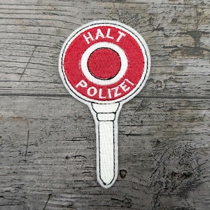 Könnte beinhalten: Gestickter Aufnäher mit einem rot-weißen Stoppschild mit den deutschen Worten "HALT POLIZEI". Der Aufnäher hat einen weißen Griff und ein kreisförmiges Design. Der Aufnäher befindet sich auf einer grauen Holzoberfläche.