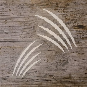 Peut inclure: Autocollants blancs en forme de griffes sur une surface en bois vieilli. Les décalcomanies sont disposées en deux groupes de trois et quatre lignes parallèles, suggérant un effet de déchirure ou de griffure. Le bois est gris et brun.