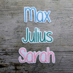 May include: Three embroidered name patches: "Max" in blue, "Julius" in teal, and "Sarah" in pink. Each name is stitched onto a white felt base with a contrasting outline. The patches are displayed on a weathered wooden surface.