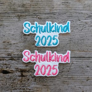 May include: Two white felt patches with the German word "Schulkind" and the year "2025" embroidered in teal and pink. The patches are on a weathered wooden surface.