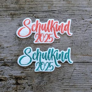 May include: Two embroidered patches with the German word "Schulkind" and the year "2025". One patch features red lettering, the other teal. Both are on a white background with a wooden background.