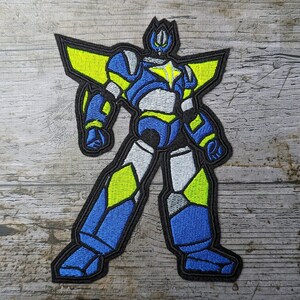 May include: Embroidered patch depicting a robot in blue, yellow, and white, with a black outline. The robot has a star on its chest and geometric shapes. Suitable for application on clothing or accessories.