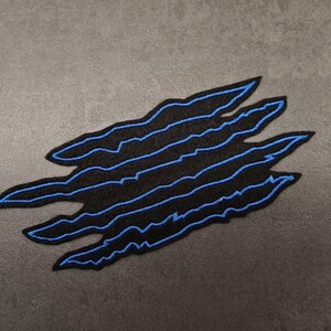 May include: Black embroidered patch featuring a claw mark design with blue stitching. The claw marks are layered and have a jagged edge. The patch is on a gray background.