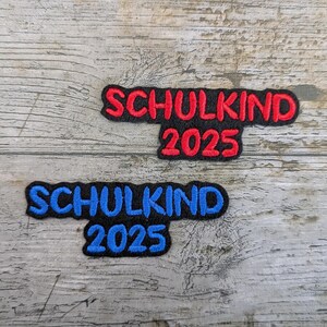 Peut inclure: Deux écussons brodés avec le texte "SCHULKIND 2025". L'un des écussons a des lettres rouges sur fond noir, l'autre a des lettres bleues sur fond noir. Les écussons sont sur une surface en bois vieilli.