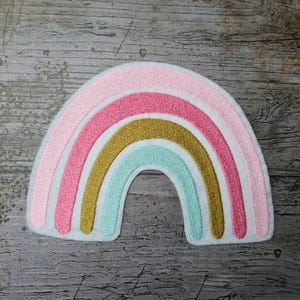 Könnte beinhalten: Gestickter Regenbogen-Aufnäher in Pastelltönen. Das Design umfasst Schichten aus Rosa, Gold und Blau, mit weißen Nähten. Dieser Aufnäher eignet sich für Kleidung, Taschen oder andere Stoffartikel.