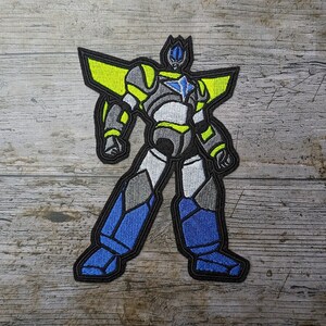 Op de afbeelding: Geborduurde patch met een robotontwerp. De robot heeft een blauwe helm, borstteken en onderbenen. De schouders en bovenarmen zijn geel, met grijze en witte accenten. De patch is zwart omlijnd.