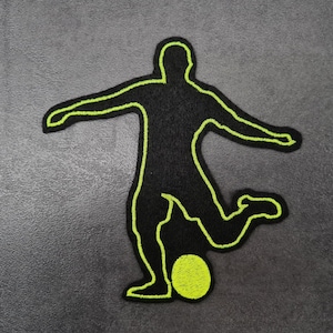 May include: Black patch featuring a soccer player silhouette kicking a ball. The player and ball are outlined in neon green against a black background. The patch is likely for sports enthusiasts.