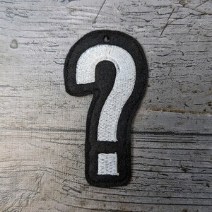 May include: A black and white embroidered question mark patch. The patch has a black border and a white question mark in the centre. It has a small hole at the top for attachment.
