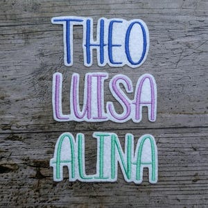 Peut inclure: Trois écussons brodés avec les noms "THEO" en bleu, "LUISA" en violet et "ALINA" en vert. Chaque nom est écrit en cursive sur fond blanc. Les écussons sont posés sur une surface en bois vieilli.