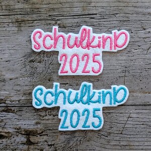 May include: Two embroidered patches with the text "Schulkind 2025". One patch has pink lettering and trim, the other has blue lettering and trim. Both patches are on a white background.