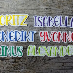 May include: Embroidered name patches in various colors. Names include Moritz, Isabella, Benedikt, Yvonne, Linus, and Alexander. Each name is stitched in a different color, with a white border, on a white background. The patches are displayed on a wooden surface.