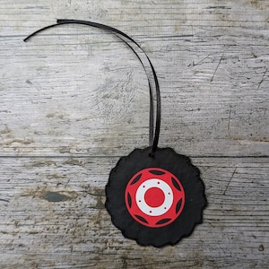 Könnte beinhalten: Ein schwarzer, kreisförmiger Schmuck in Form eines Reifens mit einem rot-weißen Nabenmuster. Der Schmuck hat einen gewellten Rand und ein schwarzes Band zum Aufhängen. Der Hintergrund ist eine verwitterte, graue Holzoberfläche.