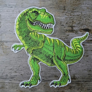 Könnte beinhalten: Gestickter grüner Tyrannosaurus Rex Aufnäher mit weißem Rand. Der Dinosaurier hat detaillierte Nähte, scharfe Zähne und gelbe Akzente. Der Aufnäher befindet sich auf einem verwitterten Holzgrund.
