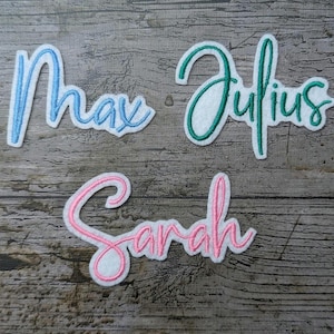 Peut inclure: Trois écussons brodés avec les noms "Max" en bleu, "Julius" en vert et "Sarah" en rose. Chaque nom est écrit en cursive avec un bord blanc, sur un fond en bois vieilli.
