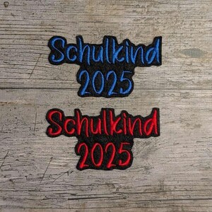 May include: Two embroidered patches with the German word "Schulkind" and the year "2025". One patch has blue text on a black background, the other has red text on a black background. The patches are on a wooden surface.