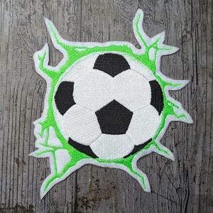 Puede incluir: Parche bordado con un diseño de balón de fútbol en blanco y negro, rodeado por un borde verde y dentado. El parche está diseñado para parecer que el balón está rompiendo una superficie. Ideal para los aficionados al deporte.