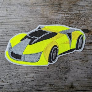 Könnte beinhalten: Gestickter Aufnäher eines gelben Sportwagens mit schwarzen und grauen Akzenten. Das Autodesign hat eine schlanke, aerodynamische Form mit detaillierten Nähten, einschließlich der Räder und Fenster. Der Aufnäher hat einen weißen Rand.