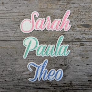 May include: Three embroidered name patches: "Sarah" in pink, "Paula" in teal, and "Theo" in blue. Each name is in a cursive script with a white background and a stitched border. The patches are arranged on a weathered, grey wooden surface.