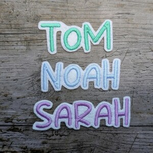 May include: Three embroidered name patches: "TOM" in green, "NOAH" in light blue, and "SARAH" in purple. Each name is stitched with a white outline, set against a wooden background.