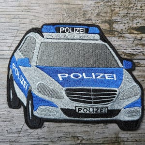 Może przedstawiać: Haftowana naszywka z radiowozu w kolorze niebieskim, szarym i białym, z napisem "POLIZEI" na samochodzie. Naszywka jest obramowana na czarno i ma szczegółowy wzór.