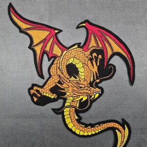 Könnte beinhalten: Gestickter Drachen-Aufnäher in Orange, Gelb und Rot mit schwarzen Umrissen. Der Drache hat ausgebreitete Flügel und einen langen, geschwungenen Schwanz. Für Kleidung oder Accessoires.
