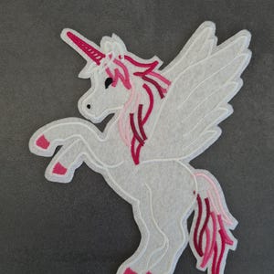 Peut inclure: Écusson de licorne blanche avec des détails roses et rouges. La licorne a une corne, une crinière et une queue roses, ainsi que des ailes blanches. L'écusson en feutre est conçu pour être cousu ou repassé sur du tissu.