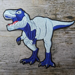 Può includere: Toppa ricamata raffigurante un dinosauro Tyrannosaurus Rex. Il design presenta un corpo azzurro con dettagli blu scuro sulla testa, le gambe e la coda. Il dinosauro ha la bocca aperta con dettagli rossi e denti bianchi affilati, delineati da un bordo nero.