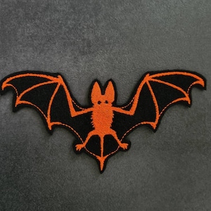 Könnte beinhalten: Gestickter Flicken in Form einer Fledermaus mit schwarzen Flügeln und einem orangefarbenen Körper. Die Fledermaus hat ausgebreitete Flügel auf grauem Hintergrund. Geeignet für Halloween- oder Gothic-Projekte.