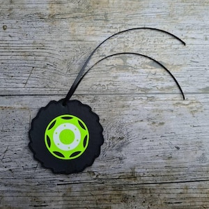 Könnte beinhalten: Ein schwarzer Monster-Truck-Reifen-förmiger Ornament mit einem neongrünen und weißen Mitteldesign. Das Ornament hat ein schwarzes Band zum Aufhängen. Das Design befindet sich auf einem Holzgrund.