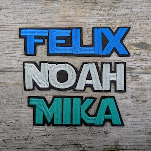 May include: Embroidered patches featuring the names FELIX, NOAH, and MIKA. FELIX is in blue, NOAH in white, and MIKA in teal. Each name is outlined in black, with a textured wood backdrop. Ideal for customising clothing or accessories.