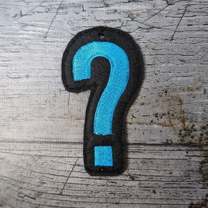 May include: A black felt question mark patch with a bright blue embroidered interior. The patch has a small hole at the top for attaching to clothing or accessories. The design is set against a weathered wood background.