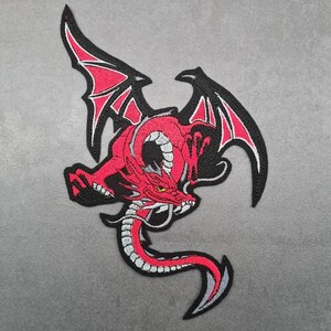 May include: Embroidered patch depicting a red dragon with black wings and a white border. The dragon has a detailed design with a long tail, set against a grey background. The patch is suitable for clothing or accessories.
