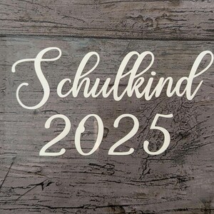 May include: White script text on a gray wooden background reads "Schulkind 2025." The text is in a cursive font, with the word "Schulkind" above the number "2025."