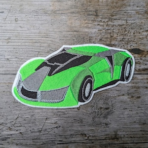 Peut inclure: Écusson brodé d'une voiture de sport verte avec des accents noirs et gris. Le design de la voiture présente une esthétique élégante et moderne avec des coutures détaillées. L'écusson a une bordure blanche et est posé sur un fond en bois.