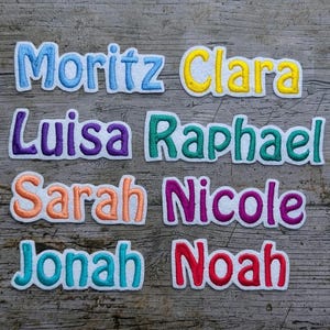 May include: Embroidered name patches in various colors, including blue, yellow, purple, green, orange, and red. Names include Moritz, Clara, Luisa, Raphael, Sarah, Nicole, Jonah, and Noah. The patches are on a wooden background.
