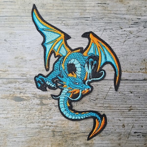 Könnte beinhalten: Gestickter Aufnäher mit einem blauen Drachen mit orangefarbenen und gelben Akzenten. Der Drache hat ausgebreitete Flügel und einen langen, geschwungenen Schwanz. Der Aufnäher ist schwarz umrandet, auf Holzgrund.