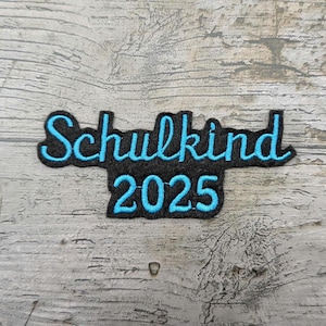 May include: A black patch with the words "Schulkind 2025" embroidered in turquoise. The text is in a cursive font. The patch is on a wooden background.